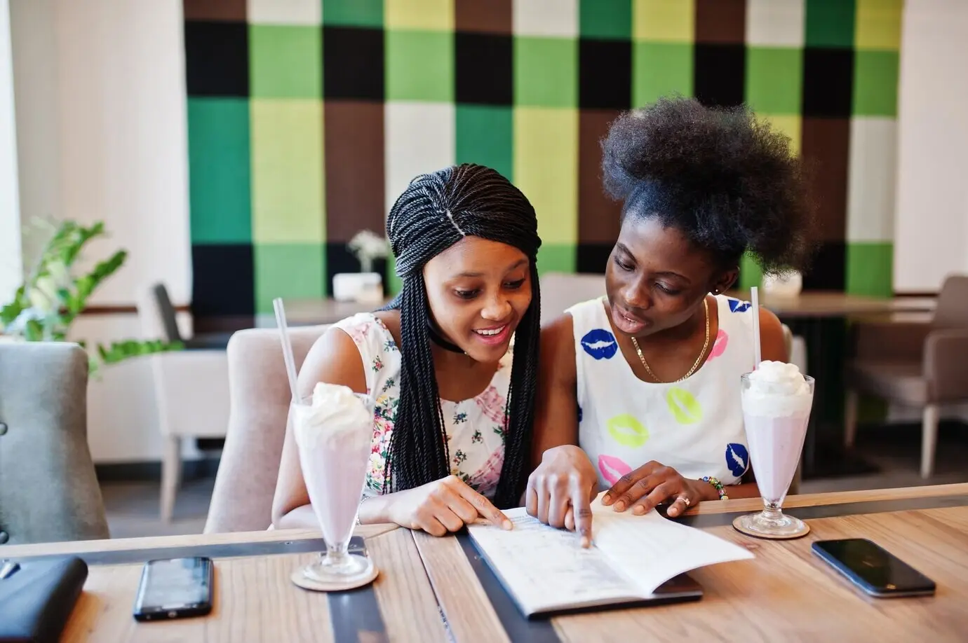 Zwei schwarze afrikanische Freundinnen in Sommerkleidern trinken Milchshake-Cocktails und schauen sich in einem Café die Speisekarte an.